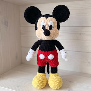 May include: A crocheted plush doll of Mickey Mouse featuring a black head and ears, a beige face, a black nose, and large white eyes. The doll has a black torso, red shorts with two white buttons, white gloves, and large yellow shoes.