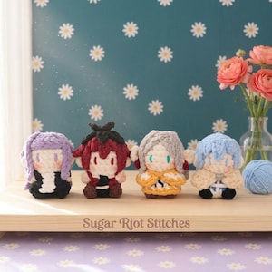 Può includere: Quattro piccole bambole all'uncinetto fatte a mano con colori di capelli e abiti distinti, disposte in fila su una tavola di legno incisa con il testo Sugar Riot Stitches. Le bambole hanno capelli in filato color lavanda, rosso, argento e blu.