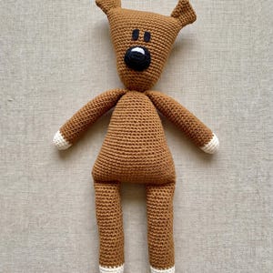 May include: A brown crocheted stuffed animal with a triangular head, two pointed ears, and a black nose with a white accent. The toy features long arms and legs, each ending in white crocheted tips, and two small black rectangular eyes.