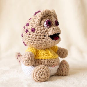 Peut inclure: Une créature en crochet rembourrée avec une peau beige, des taches violettes et de grands yeux sombres. Elle porte un haut jaune vif et une couche blanche. La figurine a une large bouche ouverte montrant une langue rouge et des dents blanches.
