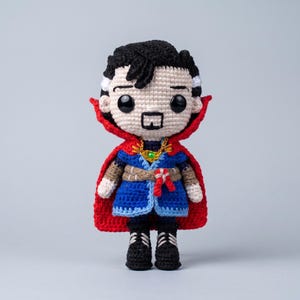 May include: A crocheted doll depicting a character with black hair, a goatee, and white streaks at the temples. The figure wears a blue tunic, a tan belt with a red knot, black boots, and a vibrant red cape featuring a gold necklace with a green gem.