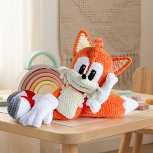 May include: A plush crochet toy of a fox character with orange and white yarn, featuring two large ears, a bushy tail, white gloves, and red and gray shoes. The character has a cream-colored muzzle, black eyes, and a small black nose.