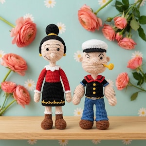 Puede incluir: Dos muñecos de ganchillo hechos a mano que representan a un personaje femenino con moño negro, top rojo y falda negra, y a un personaje masculino con gorro de marinero blanco, camisa negra, pantalones azules y una pipa amarilla en la boca.