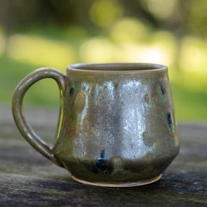 May include: A handmade ceramic mug with a rounded body and a curved handle. The mug has a mottled glaze in shades of green, brown, and blue. The mug is sitting on a wooden surface, with a blurred green background.