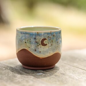 May include: A handmade ceramic cup with a brown base and a blue and tan speckled upper section. A crescent moon is carved into the blue section. The cup is approximately 7.6 cm tall and 7.6 cm wide.