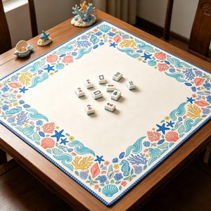 Könnte beinhalten: Eine cremefarbene Mahjong-Spielmatte mit blauem Rand und einem farbenfrohen Design zum Thema Ozean. Der Rand zeigt Illustrationen von Muscheln, Seesternen und Korallen. Mehrere Mahjong-Steine sind auf der Matte verstreut.