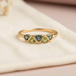 Puede incluir: Un anillo de oro con tres piedras preciosas triangulares. El anillo tiene una banda delgada y está adornado con piedras azules y verdes. El anillo se muestra sobre una tela blanca, con un fondo rosa claro.