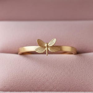 May include: A gold ring with a dragonfly motif. The dragonfly is the central feature, with intricate wing detailing. The ring is presented in a pink jewellery box, accentuating the ring's delicate design.