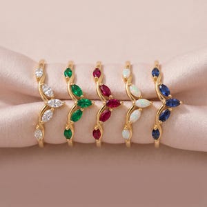 Puede incluir: Una colección de anillos dorados, cada uno con una disposición única de piedras preciosas. Los anillos muestran un diseño en forma de hoja con piedras blancas, verdes, rojas, ópalo y azules. Los anillos se exhiben sobre una tela rosa.