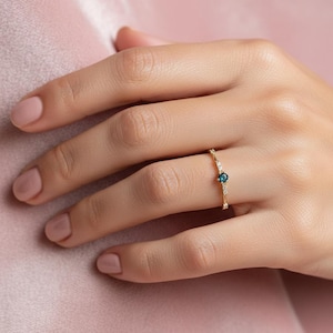 Puede incluir: Un delicado anillo de oro con una pequeña gema redonda de color azul oscuro en el centro, acentuada por piedras más pequeñas y transparentes. El anillo está en un dedo, sobre un fondo rosa suave.