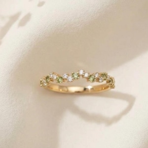 May include: A gold ring with a delicate design. The ring features small, round, white and green gemstones set along the band. The gemstones are arranged in a flowing pattern. The ring is set against a soft, off-white background.