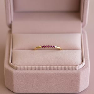 Peut inclure: Une bague en or ornée d'une rangée de petites pierres précieuses rouges. La bague est présentée dans un écrin à bijoux rose doublé de velours, sur un fond flou.