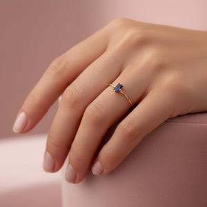 May include: A gold ring with a rectangular, dark blue gemstone. The ring is delicate and sits on a finger. The background is a soft pink color.
