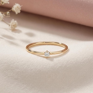 Puede incluir: Un delicado anillo de oro con una sola gema redonda y transparente en el centro. La banda del anillo es fina y pulida, reflejando la luz. El anillo está sobre un fondo de tela suave y blanquecina.