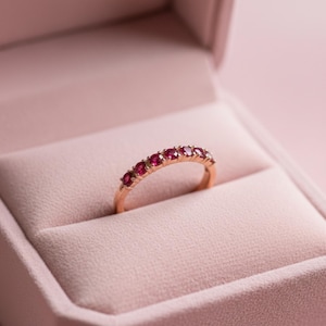 Puede incluir: Un anillo de oro con una fila de siete piedras preciosas rojas. El anillo se presenta en un joyero rosa, creando una estética suave y romántica. Las piedras preciosas están espaciadas uniformemente y capturan la luz.