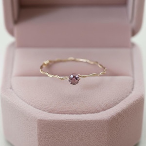 Puede incluir: Un delicado anillo de oro con una banda trenzada y una piedra preciosa redonda de color púrpura claro. El anillo se exhibe dentro de un joyero de terciopelo rosa, creando una presentación suave y elegante. El diseño del anillo es simple pero sofisticado.