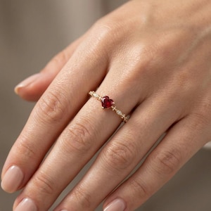 May include: A gold ring featuring a heart-shaped, deep red gemstone as the centerpiece. The band is adorned with small, sparkling accents. The ring is a delicate piece of jewelry, perfect for a romantic occasion.