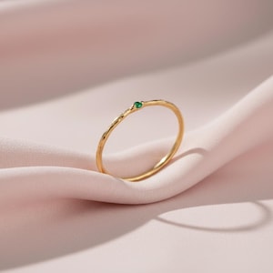 Puede incluir: Un delicado anillo de oro con una banda texturizada y una pequeña gema verde redonda. El anillo se presenta sobre un fondo de tela rosa suave, creando una estética minimalista y elegante. Es una joya.