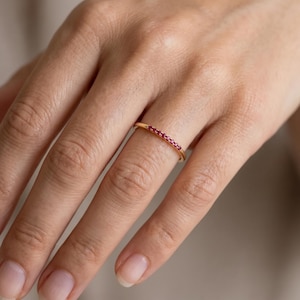 Peut inclure: Une fine bague en or ornée d'une rangée de petites pierres précieuses rouges vives. La bague est mise en valeur sur un fond neutre, soulignant le design élégant de la bague et la couleur riche des pierres. La bague est de style simple.
