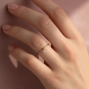 May include: A delicate gold ring with a small, dangling diamond pendant. The ring is thin and minimalist, with a simple band and a tiny, sparkling gemstone. The ring is shown on a person's finger, highlighting its elegant design.