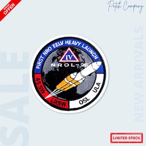 May include: Embroidered patch featuring a rocket launch design with the text "FIRST NRO EELV HEAVY LAUNCH". The patch includes the text "NROL-26", "45 SW", "LRSW", and "OSL". The patch has a blue, white, yellow, and black color scheme.