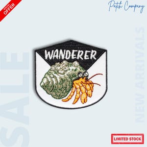 May include: Embroidered patch featuring a hermit crab with the word "WANDERER" in white on a black and white shield design. The crab is green and orange. The patch is a special offer and limited stock.