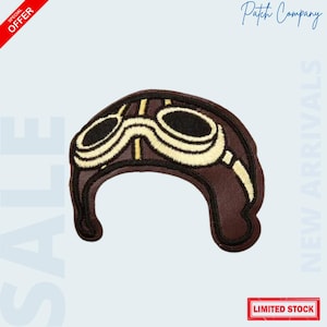 May include: A brown embroidered patch in the shape of a vintage aviator helmet with goggles. The goggles are cream-colored with black lenses. The patch has a black border and the text "Patch Company" is visible.