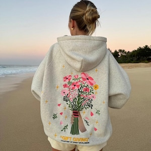Puede incluir: Una sudadera con capucha gris claro con un bordado de ramo floral en la espalda. El ramo presenta flores rosas y amarillas con tallos verdes. El texto "GIFT GIVING" está debajo. La sudadera con capucha se usa en una playa.