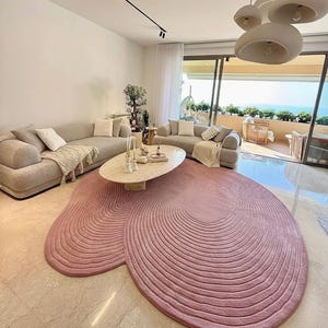 May include: A large, irregular, dusty pink area rug featuring a textured, concentric line pattern that mimics organic, fluid shapes. The rug is positioned on a light-colored floor in a living space, partially underneath an oval-shaped coffee table.