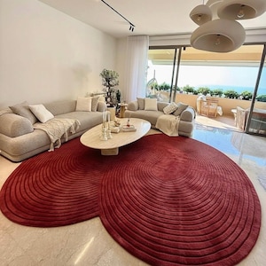 May include: A large, deep red area rug with a textured, concentric circular pattern that forms an irregular, organic shape. The rug is positioned on a light-colored marble floor in a living room, extending beneath an oval coffee table and sofas.
