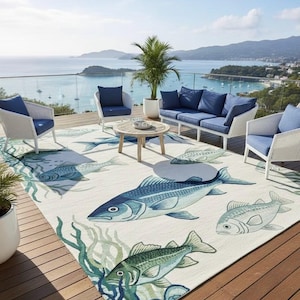 Puede incluir: Una alfombra de patio exterior con temática marina. La alfombra presenta ilustraciones grandes y detalladas de peces en tonos azules y verdes sobre un fondo crema. Muebles de mimbre blanco con cojines azules rodean la alfombra, con vistas a la costa.