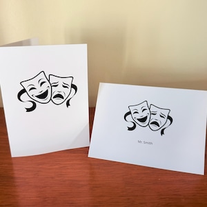 May include: Two white folded greeting cards featuring a black graphic of the comedy and tragedy theater masks. One card is standing vertically, while the other is placed horizontally in front, displaying the printed text Mr. Smith below the masks.