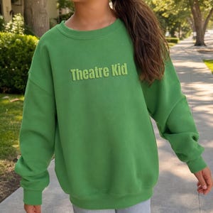 May include: A bright green crewneck sweatshirt featuring the text Theatre Kid in raised, matching green lettering across the chest. The garment has long sleeves with ribbed cuffs and a ribbed neckline, providing a simple and uniform appearance.