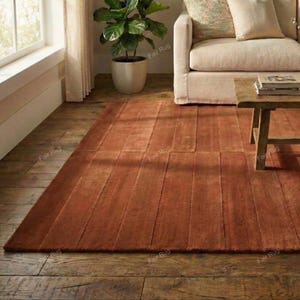 Puede incluir: Una alfombra rectangular de color óxido con un diseño texturizado y acanalado. La alfombra está colocada sobre un suelo de madera y la habitación presenta una paleta de colores neutros con un sofá, una mesa de centro y una planta en maceta.