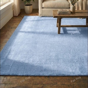 Könnte beinhalten: Ein hellblaues rechteckiges Rug mit einer weichen, strukturierten Oberfläche. Der Teppich liegt auf einem Holzboden, mit einem Couchtisch und einem beigen Sofa im Hintergrund. Der Teppich hat einen dezenten Rand.