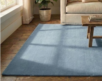 Puede incluir: Una alfombra rectangular azul claro sobre un suelo de madera. La alfombra está en un salón, con un sofá beige, una mesa de centro de madera y una planta en maceta en el fondo. La alfombra es de color liso y parece estar hecha de un material suave.