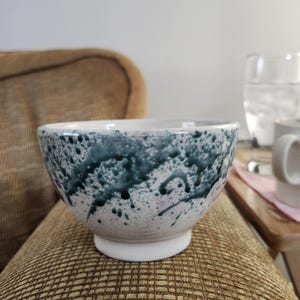 May include: A ceramic bowl with a white base and a speckled design in shades of teal and white. The bowl has a rounded shape and is sitting on a brown patterned surface.