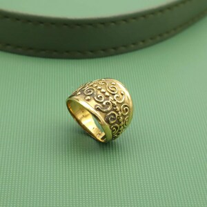 Può includere: Un anello color oro con un motivo a spirale ornato. L'anello ha una forma arrotondata e una finitura lucida. Il design presenta volute dettagliate e motivi floreali, creando un'estetica vintage. L'anello è posto su uno sfondo verde.