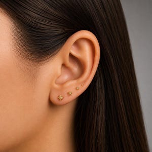 Pode incluir: Um conjunto de quatro brincos dourados dispostos em fila ao longo do lóbulo da orelha. Cada brinco apresenta um pequeno design de estrela de várias pontas com um ponto central, diminuindo de tamanho da maior estrela para o menor ponto.