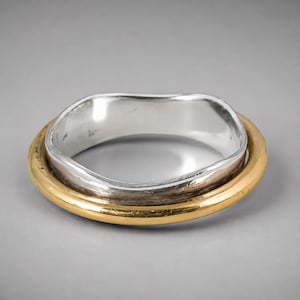 May include: A two-tone ring featuring an inner band with a wavy, irregular silver-colored edge and a smooth, circular outer band with a polished gold-colored finish. The design consists of two distinct metallic rings nested together.
