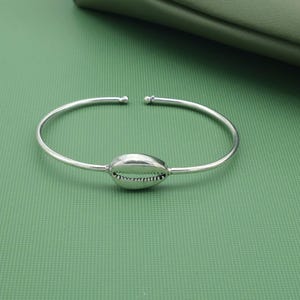 May include: A silver-colored cuff bracelet featuring a seashell design at the center. The bracelet has an open design with rounded ends. The seashell is detailed with textured ridges and a smooth, polished surface.