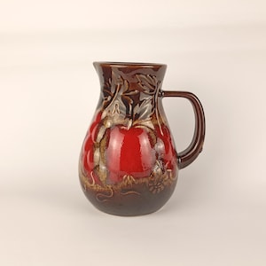 Puede incluir: Una jarra de cerámica con acabado brillante, base marrón oscuro y centro rojo vibrante. La superficie está decorada con un patrón en relieve de manzanas y hojas que rodea el cuerpo del recipiente.