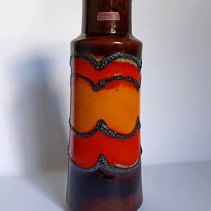 Può includere: Un vaso alto in ceramica con una combinazione di colori marrone, arancione e rosso. Il vaso ha una forma cilindrica con una base svasata e un collo stretto. Il design include fasce ondulate e strutturate in colori contrastanti. La parola "Scheurich" è visibile su una piccola etichetta.