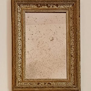Può includere: Uno specchio da parete rettangolare con una cornice color oro invecchiato e intricati motivi intagliati. La superficie del vetro presenta una distinta patina invecchiata e maculata con punti scuri, inserita in un bordo a più livelli.