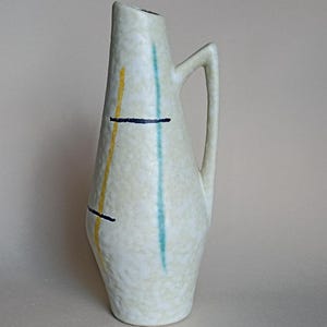 Può includere: Brocca alta in ceramica bianco sporco con manico. La brocca è decorata con strisce verticali gialle e turchesi e linee nere orizzontali. Ha una superficie strutturata e un'apertura stretta.