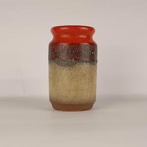 Puede incluir: Un jarrón cilíndrico de cerámica con un borde esmaltado de color rojo vibrante que transiciona hacia una banda marrón oscura y texturizada, seguida de una base beige moteada. El recipiente tiene una abertura redondeada y un cuerpo recto.