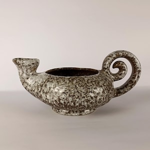 Può includere: Una lampada a olio in ceramica con una smaltatura maculata marrone e bianca. Il recipiente presenta un corpo largo e arrotondato, un beccuccio corto su un lato e un manico prominente e arricciato sul lato opposto. L'interno è marrone scuro.