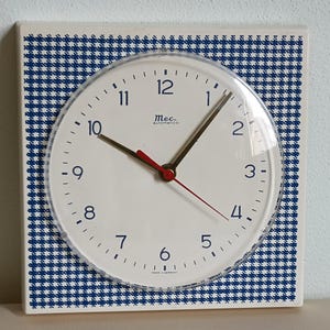 Può includere: Un orologio da parete bianco con una copertura in plastica trasparente, numeri e lancette nere e una lancetta dei secondi rossa. Il quadrante dell'orologio ha il testo "Mec automatico". La cornice quadrata ha un motivo a scacchi blu e bianco. Made in Germany.