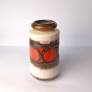 Può includere: Un vaso cilindrico in ceramica con un bordo marrone spesso, un corpo color crema e una fascia centrale di vivaci motivi circolari arancioni su uno sfondo marrone testurizzato. È visibile una piccola etichetta rettangolare con la scritta Scheurich.