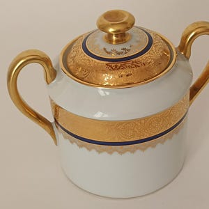 Puede incluir: Azucarero de porcelana blanca con tapa y asas doradas. La tapa y una banda alrededor del cuenco presentan intrincados diseños de filigrana dorada y un acento azul marino. El azucarero tiene un pomo dorado en la parte superior.